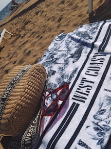 On a sandy beach, a patterned towel with the text 'WEST COAST' is spread out. A pair of red sunglasses and a woven bag with straps are placed on the towel. The ocean is visible in the background with some people and beach chairs near the shore.