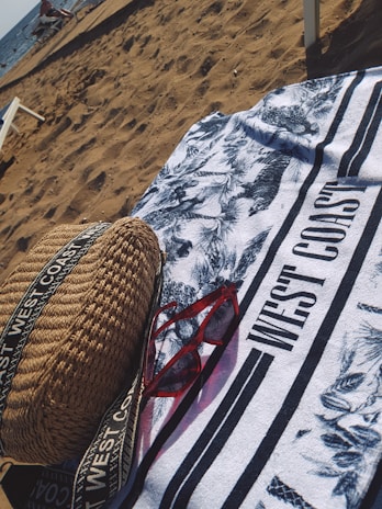 On a sandy beach, a patterned towel with the text 'WEST COAST' is spread out. A pair of red sunglasses and a woven bag with straps are placed on the towel. The ocean is visible in the background with some people and beach chairs near the shore.