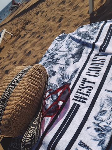 On a sandy beach, a patterned towel with the text 'WEST COAST' is spread out. A pair of red sunglasses and a woven bag with straps are placed on the towel. The ocean is visible in the background with some people and beach chairs near the shore.