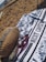 On a sandy beach, a patterned towel with the text 'WEST COAST' is spread out. A pair of red sunglasses and a woven bag with straps are placed on the towel. The ocean is visible in the background with some people and beach chairs near the shore.