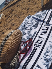 On a sandy beach, a patterned towel with the text 'WEST COAST' is spread out. A pair of red sunglasses and a woven bag with straps are placed on the towel. The ocean is visible in the background with some people and beach chairs near the shore.