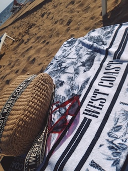 On a sandy beach, a patterned towel with the text 'WEST COAST' is spread out. A pair of red sunglasses and a woven bag with straps are placed on the towel. The ocean is visible in the background with some people and beach chairs near the shore.