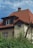 A house with a red tiled roof, featuring a chimney and dormer windows, surrounded by lush green foliage with some red berries visible. The walls are cream-colored, and the sky above is partly cloudy.