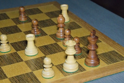 A close-up view of a chessboard featuring several chess pieces in the middle of a game. The pieces are made of wood, with a mix of light and dark tones. The board has a classic checkered pattern and is positioned on a blue surface.