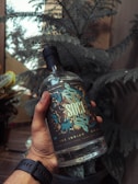 A hand is holding a bottle with intricate and colorful label design, against a backdrop of green foliage. The bottle appears to be of gin, indicated by the labeling. The overall setting is indoors, with natural light streaming in from a window.