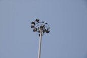 A tall metal pole featuring a circular arrangement of mounted floodlights against a clear blue sky. The lights are arranged symmetrically at the top of the pole.
