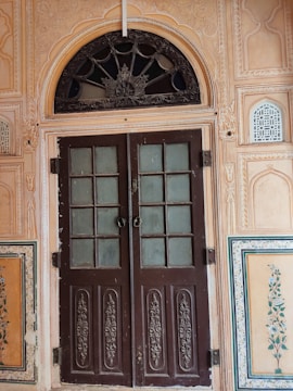 An intricately carved wooden door with two panels stands in the middle. The door features frosted glass windows and ornate designs on the lower sections. Surrounding the door is a decorative frame with detailed floral and geometric patterns. The arched transom above the door includes a decorative metal grille with floral motifs. The walls are adorned with paintings of floral designs and have textured and patterned surfaces.
