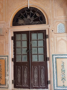 An intricately carved wooden door with two panels stands in the middle. The door features frosted glass windows and ornate designs on the lower sections. Surrounding the door is a decorative frame with detailed floral and geometric patterns. The arched transom above the door includes a decorative metal grille with floral motifs. The walls are adorned with paintings of floral designs and have textured and patterned surfaces.