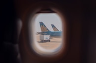 The view from an airplane window shows two aircraft tails with a floral logo parked on the tarmac, along with a fuel truck. The scene is framed through the rounded window, capturing a sense of travel and transportation.