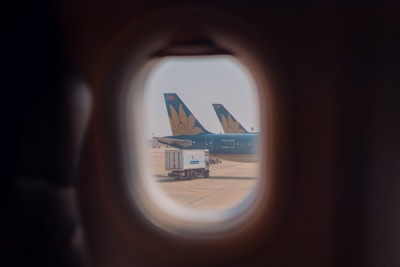 The view from an airplane window shows two aircraft tails with a floral logo parked on the tarmac, along with a fuel truck. The scene is framed through the rounded window, capturing a sense of travel and transportation.