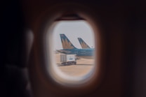 The view from an airplane window shows two aircraft tails with a floral logo parked on the tarmac, along with a fuel truck. The scene is framed through the rounded window, capturing a sense of travel and transportation.