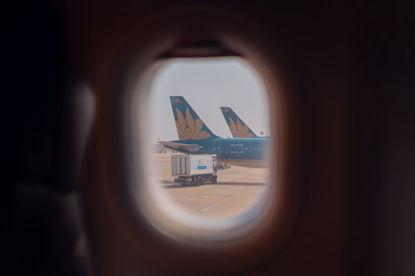 The view from an airplane window shows two aircraft tails with a floral logo parked on the tarmac, along with a fuel truck. The scene is framed through the rounded window, capturing a sense of travel and transportation.