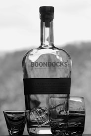 A large glass bottle with the label 'Boondocks American Whiskey' stands prominently in the center, accompanied by two empty whiskey glasses and a shot glass placed in front of it. The background is softly blurred, focusing attention on the bottle and glasses.