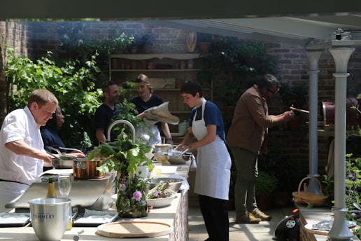 A group of people are engaged in cooking, surrounded by green plants and brick walls. Some are preparing food at a counter with pots and ingredients, while one person is operating a rotating grill. The atmosphere is casual and cooperative, with sunlight filtering through the space.