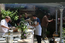 A group of people are engaged in cooking, surrounded by green plants and brick walls. Some are preparing food at a counter with pots and ingredients, while one person is operating a rotating grill. The atmosphere is casual and cooperative, with sunlight filtering through the space.