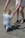 A small child in a blue and white striped outfit is being supported while walking on a sandy beach, holding onto the hands of an adult wearing white shorts and black sandals.