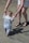 A small child in a blue and white striped outfit is being supported while walking on a sandy beach, holding onto the hands of an adult wearing white shorts and black sandals.