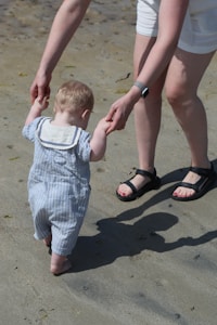 A small child in a blue and white striped outfit is being supported while walking on a sandy beach, holding onto the hands of an adult wearing white shorts and black sandals.