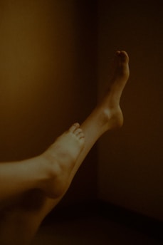 A pair of legs are extended against a warmly lit, plain background. The lighting creates a soft, shadowy effect around the legs, emphasizing curves and contours.