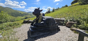 A black stone statue of a cow with intricate carvings sits on a raised platform in a scenic outdoor setting. The background features lush green hills under a vivid blue sky with scattered white clouds. The area surrounding the statue is covered in gravel, with some large rocks placed around. The setting indicates a serene and peaceful environment.