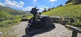 A black stone statue of a cow with intricate carvings sits on a raised platform in a scenic outdoor setting. The background features lush green hills under a vivid blue sky with scattered white clouds. The area surrounding the statue is covered in gravel, with some large rocks placed around. The setting indicates a serene and peaceful environment.