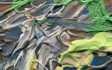 A textured abstract painting featuring swirling patterns of paint in shades of green, blue, gray, and brown. The paint appears to be thick and possibly applied with a palette knife, creating a dynamic and organic composition.