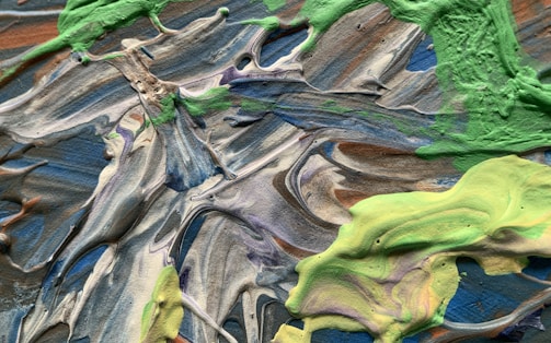A textured abstract painting featuring swirling patterns of paint in shades of green, blue, gray, and brown. The paint appears to be thick and possibly applied with a palette knife, creating a dynamic and organic composition.