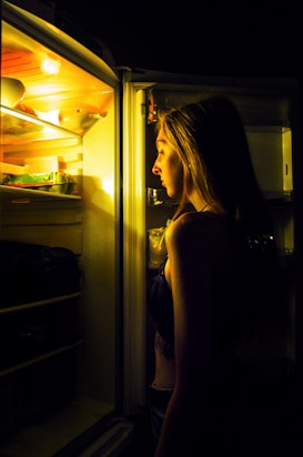 A woman is standing in front of an open refrigerator, illuminated by the warm yellow light from inside the fridge. The scene takes place in a dark room, creating a stark contrast between the light from the fridge and the surrounding darkness.