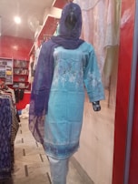 A mannequin is dressed in a light blue embroidered kurta with white floral patterns, paired with a matching blue dupatta. The mannequin is displayed in a clothing store with a red and white interior. Various fabrics and garments are visible in the background.