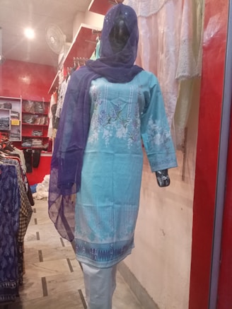 A mannequin is dressed in a light blue embroidered kurta with white floral patterns, paired with a matching blue dupatta. The mannequin is displayed in a clothing store with a red and white interior. Various fabrics and garments are visible in the background.