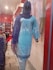 A mannequin is dressed in a light blue embroidered kurta with white floral patterns, paired with a matching blue dupatta. The mannequin is displayed in a clothing store with a red and white interior. Various fabrics and garments are visible in the background.