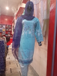 A mannequin is dressed in a light blue embroidered kurta with white floral patterns, paired with a matching blue dupatta. The mannequin is displayed in a clothing store with a red and white interior. Various fabrics and garments are visible in the background.