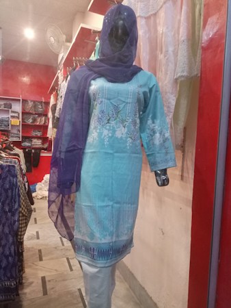 A mannequin is dressed in a light blue embroidered kurta with white floral patterns, paired with a matching blue dupatta. The mannequin is displayed in a clothing store with a red and white interior. Various fabrics and garments are visible in the background.