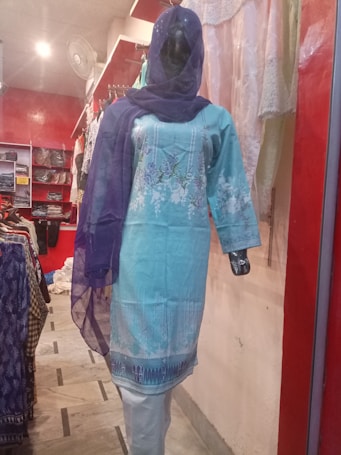 A mannequin is dressed in a light blue embroidered kurta with white floral patterns, paired with a matching blue dupatta. The mannequin is displayed in a clothing store with a red and white interior. Various fabrics and garments are visible in the background.