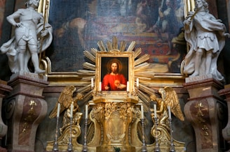 A detailed religious altar is adorned with a central painting depicting a figure in red with a serene expression, surrounded by ornate golden decorations and two angel statues. Flanking the altar are two white statues of saints labeled S. Mauritus and S. Amadeus. The background features an intricate mural.