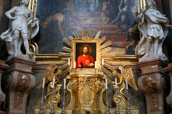 A detailed religious altar is adorned with a central painting depicting a figure in red with a serene expression, surrounded by ornate golden decorations and two angel statues. Flanking the altar are two white statues of saints labeled S. Mauritus and S. Amadeus. The background features an intricate mural.