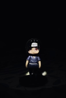 A small figurine with black hair and red eyes, wearing a dark outfit and a headband with a symbol, stands against a stark, dark background. A beam of light highlights the figure, creating a dramatic contrast with the surrounding darkness.