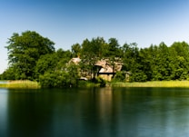 A tranquil lake scene with calm water reflecting the vibrant green of the surrounding trees. A house with a sloped roof partially obscured by the foliage stands on the shoreline, creating a sense of seclusion and harmony with nature. The sky is clear, with a gradient from blue to lighter shades as it meets the horizon.