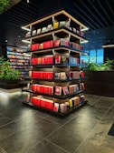 A modern library or bookstore interior features a prominently displayed multi-tiered shelf filled with neatly arranged books, predominantly with red spines. The shelves are accented by soft glowing lights, and the surrounding area has additional bookshelves and some greenery, contributing to a contemporary and inviting atmosphere. The floor is made of dark tiles, complementing the dark tones of the ceiling.