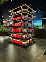 A modern library or bookstore interior features a prominently displayed multi-tiered shelf filled with neatly arranged books, predominantly with red spines. The shelves are accented by soft glowing lights, and the surrounding area has additional bookshelves and some greenery, contributing to a contemporary and inviting atmosphere. The floor is made of dark tiles, complementing the dark tones of the ceiling.
