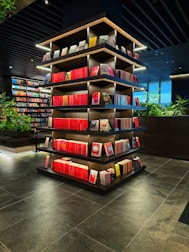 A modern library or bookstore interior features a prominently displayed multi-tiered shelf filled with neatly arranged books, predominantly with red spines. The shelves are accented by soft glowing lights, and the surrounding area has additional bookshelves and some greenery, contributing to a contemporary and inviting atmosphere. The floor is made of dark tiles, complementing the dark tones of the ceiling.