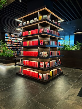 A modern library or bookstore interior features a prominently displayed multi-tiered shelf filled with neatly arranged books, predominantly with red spines. The shelves are accented by soft glowing lights, and the surrounding area has additional bookshelves and some greenery, contributing to a contemporary and inviting atmosphere. The floor is made of dark tiles, complementing the dark tones of the ceiling.
