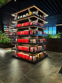 A modern library or bookstore interior features a prominently displayed multi-tiered shelf filled with neatly arranged books, predominantly with red spines. The shelves are accented by soft glowing lights, and the surrounding area has additional bookshelves and some greenery, contributing to a contemporary and inviting atmosphere. The floor is made of dark tiles, complementing the dark tones of the ceiling.