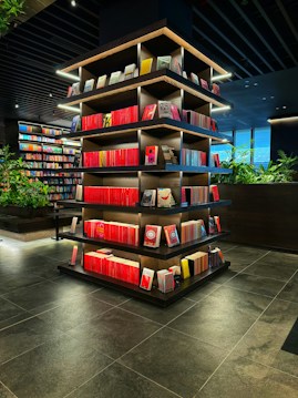 A modern library or bookstore interior features a prominently displayed multi-tiered shelf filled with neatly arranged books, predominantly with red spines. The shelves are accented by soft glowing lights, and the surrounding area has additional bookshelves and some greenery, contributing to a contemporary and inviting atmosphere. The floor is made of dark tiles, complementing the dark tones of the ceiling.