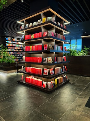 A modern library or bookstore interior features a prominently displayed multi-tiered shelf filled with neatly arranged books, predominantly with red spines. The shelves are accented by soft glowing lights, and the surrounding area has additional bookshelves and some greenery, contributing to a contemporary and inviting atmosphere. The floor is made of dark tiles, complementing the dark tones of the ceiling.