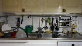 A kitchen countertop with a double sink is cluttered with various kitchen items. On the left, clean plates are drying on a dish rack. Above, there are cabinets and a white tiled wall with kitchen tools like scissors, a peeler, and a ladle hanging. A green watering can is placed next to a dishwashing sponge on the sink. To the right, there are bottles of oil and vinegar along with some garlic.