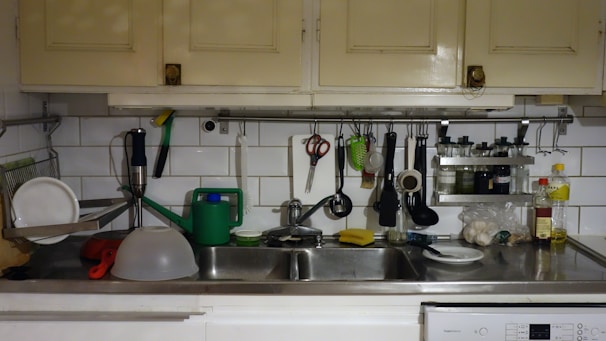 A kitchen countertop with a double sink is cluttered with various kitchen items. On the left, clean plates are drying on a dish rack. Above, there are cabinets and a white tiled wall with kitchen tools like scissors, a peeler, and a ladle hanging. A green watering can is placed next to a dishwashing sponge on the sink. To the right, there are bottles of oil and vinegar along with some garlic.
