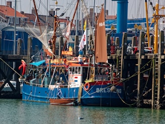 A vibrant and colorful fishing boat is docked at a busy harbor, showcasing a mix of traditional and modern maritime elements. The boat is adorned with nautical flags and fishing equipment. Surrounding it are other maritime vessels and structures made of wood and metal, with buildings and cranes visible in the background.