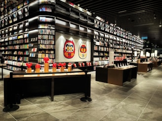 A modern bookstore interior featuring large, dark shelves filled with books and displayed art, including traditional Japanese Daruma dolls. The space is well-lit and organized, with tables showcasing various items and decorations.