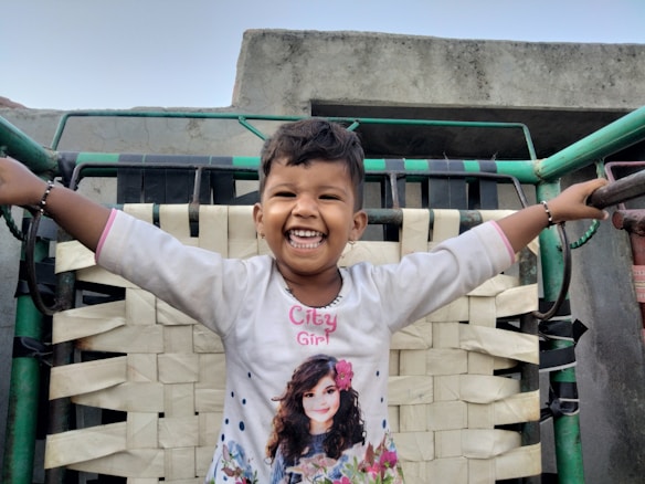 A young child is joyfully smiling, standing with arms outstretched on a woven cot with a backdrop of a concrete structure. The child is wearing a white shirt with a graphic design, featuring a girl with flowers.